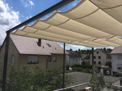 Zwei hellbeige Faltsonnensegel spenden Schatten über einer Terrasse, dahinter stehen Wohnhäuser mit Ziegeldächern unter blauem, leicht bewölktem Himmel.